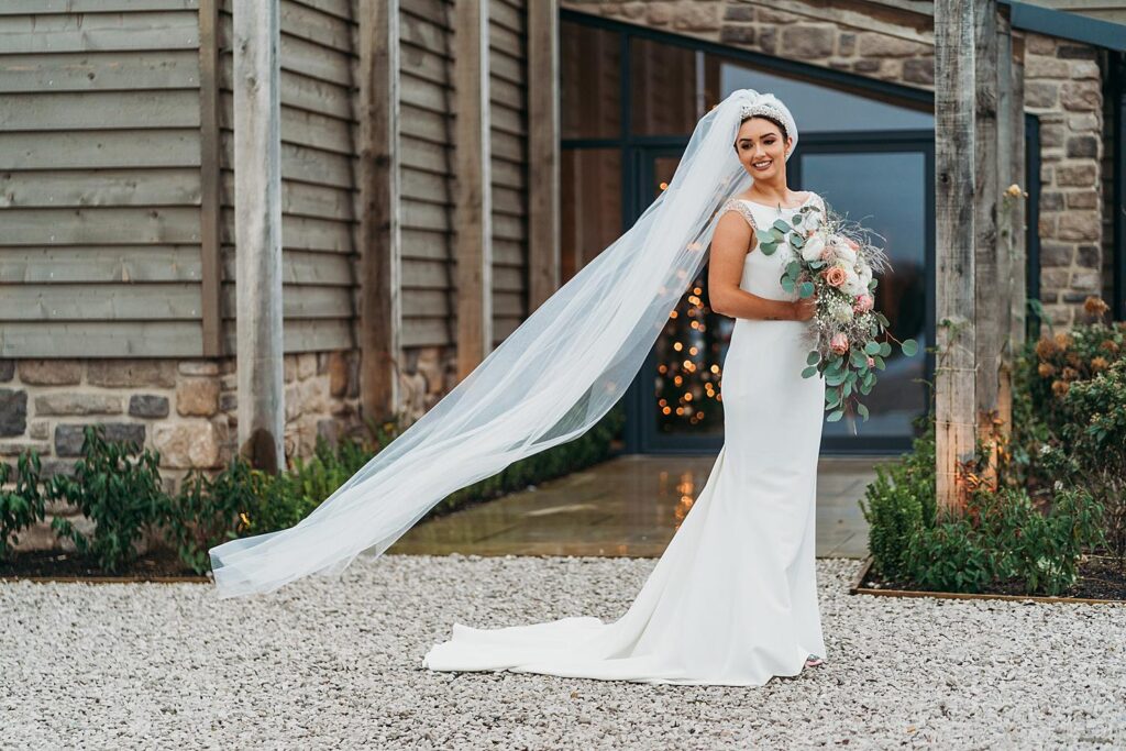 Beautiful bride stood outside Stretton Manor Barn holding bouquet whilst veil flows behind her